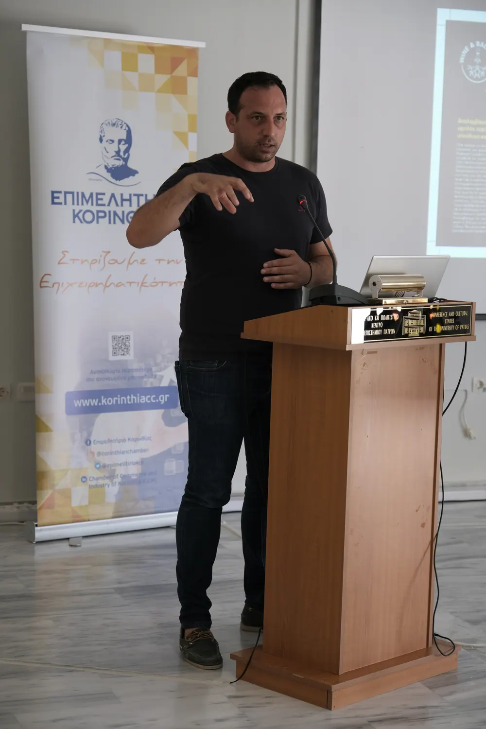 Ομιλητής στο βήμα της εκδήλωσης Wine & Balance στο Πανεπιστήμιο Πατρών, με το roll-up του Επιμελητηρίου Κορινθίας δίπλα του.