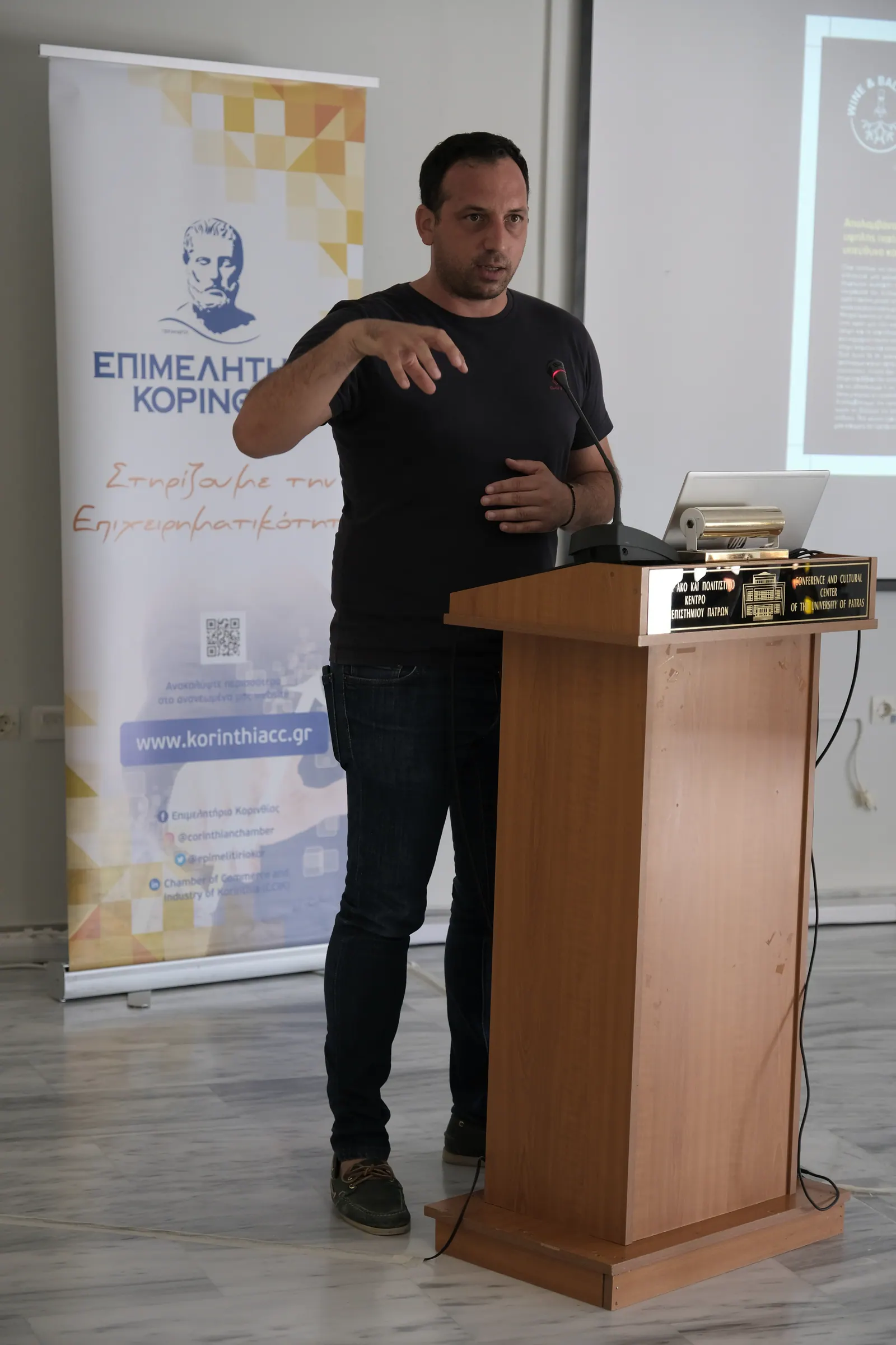 Ομιλητής στο βήμα της εκδήλωσης Wine & Balance στο Πανεπιστήμιο Πατρών, με το roll-up του Επιμελητηρίου Κορινθίας δίπλα του.